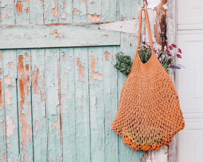 A mesh market bag full of vegetables, fruits, and flowers hanging on a green door's hinge.