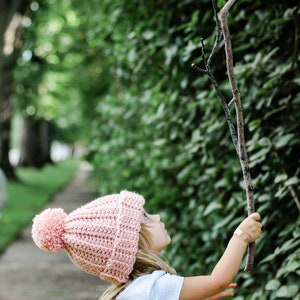 A little blonde girl wearing a pink beginner child hat holding a tree branch.