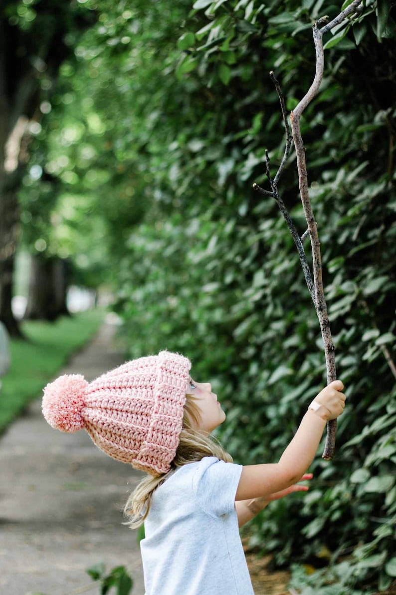 A little blonde girl wearing a pink beginner child hat holding a tree branch.