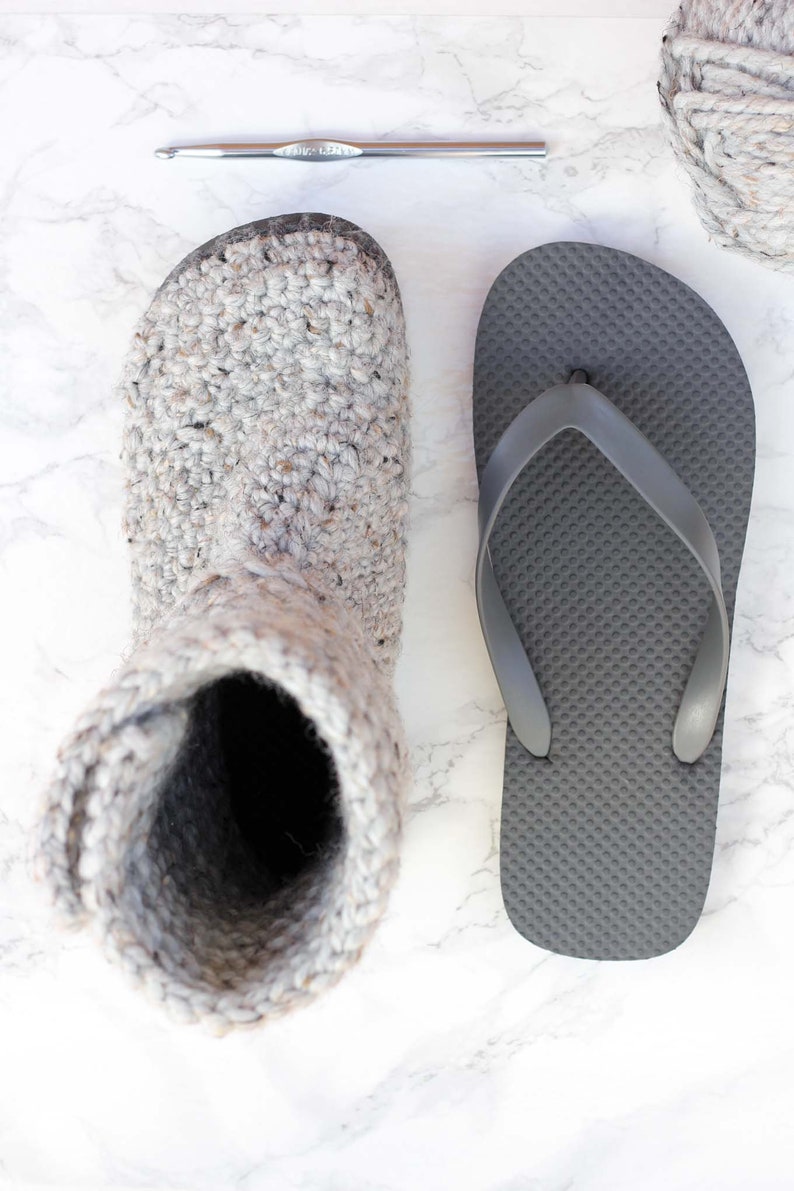 A crochet shoe with flip-flop soles, a gray flip-flop, a crochet hook, and a yarn skein.