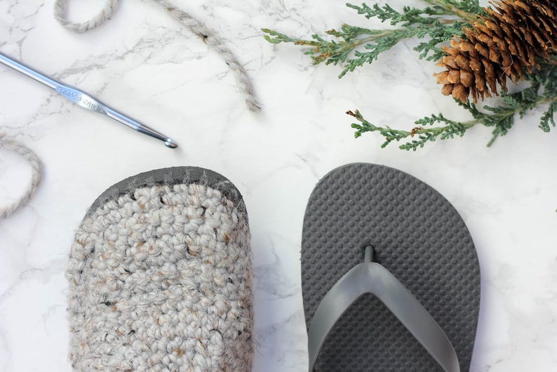 A crochet shoe with flip-flop soles, a gray flip-flop, a crochet hook, and a decorative pinecone.