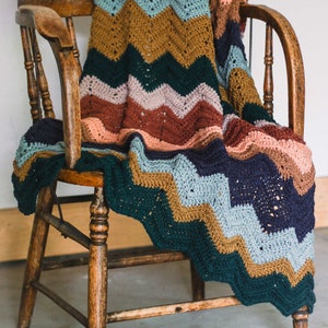 May include: A multi-colored crocheted blanket with a chevron pattern draped over a wooden chair. The blanket features shades of green, blue, brown, pink, and peach.