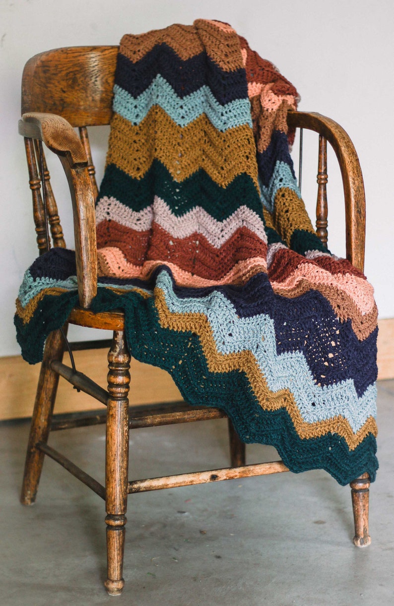 May include: A multi-colored crocheted blanket with a chevron pattern draped over a wooden chair. The blanket features shades of green, blue, brown, pink, and peach.