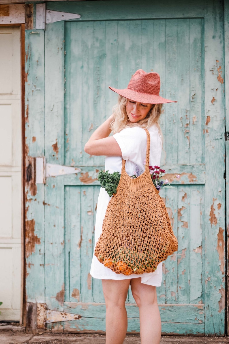 A blonde woman standing in front of a green door, looking down, wearing a sun hat and a white dress, with an easy tote bag full of vegetables, fruits, and flowers.