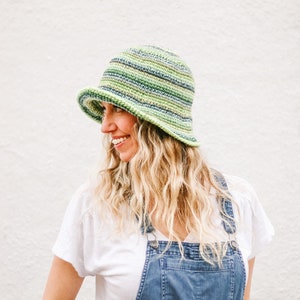 A blonde woman stands in front of a white wall, smiling, looking sideways while wearing a green bucket hat, a white T-shirt, and denim overalls.