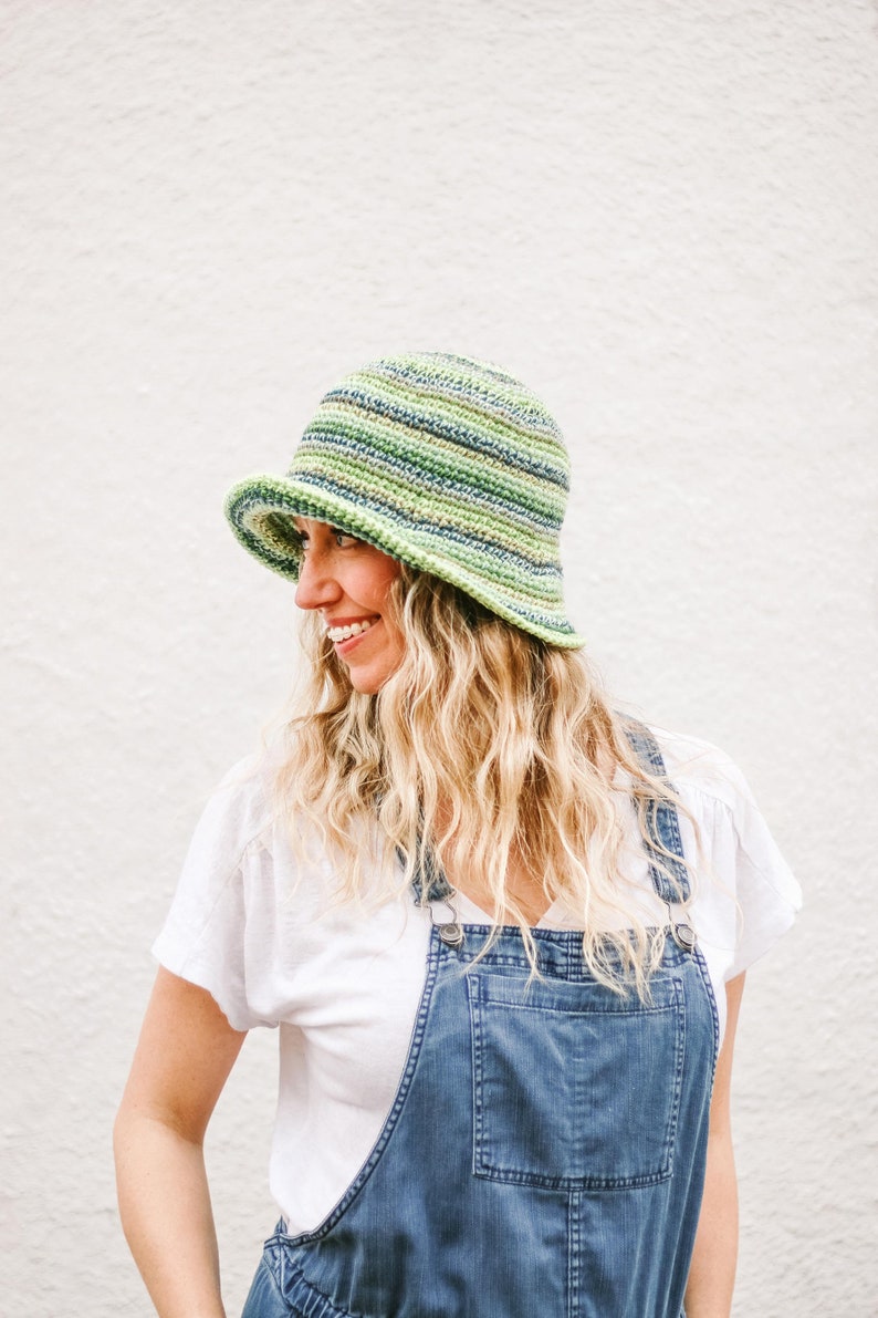 A blonde woman stands in front of a white wall, smiling, looking sideways while wearing a green bucket hat, a white T-shirt, and denim overalls.