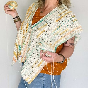 A blonde woman with a half-finished crochet top on her torso. She is holding a ball of yarn in each hand and smiling.