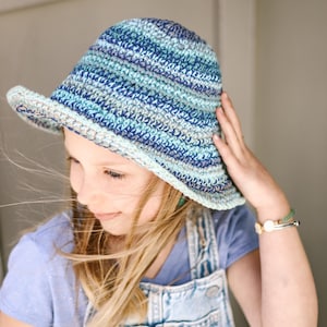 A little blonde girl smiling, wearing a blue bucket hat, a lilac t-shirt, and a denim jumper with her hand on the hat.