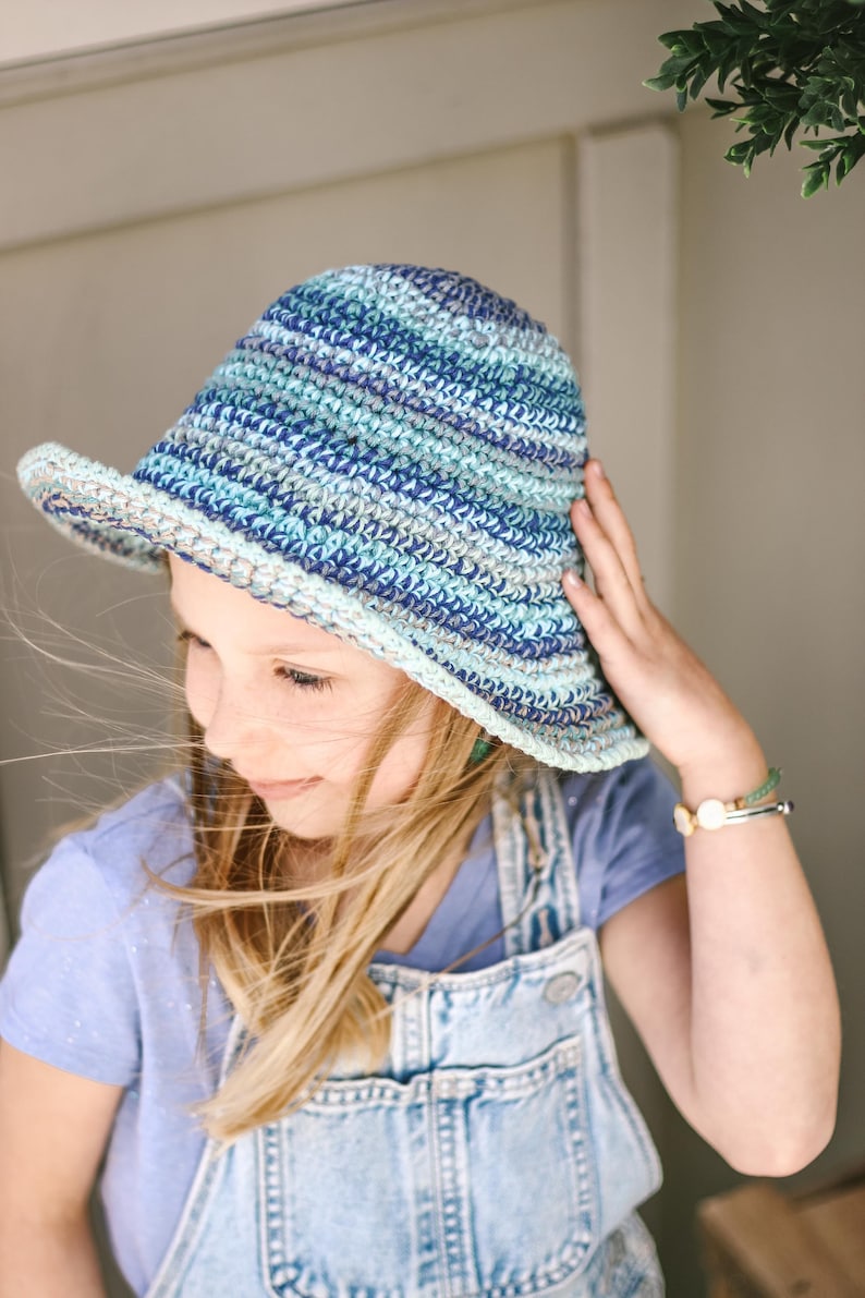 A little blonde girl smiling, wearing a blue bucket hat, a lilac t-shirt, and a denim jumper with her hand on the hat.