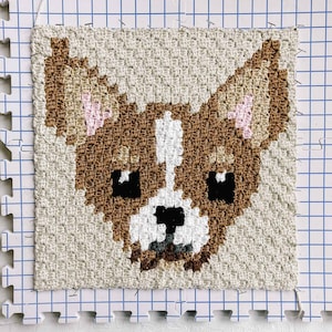May include: A brown and white chihuahua dog is depicted in a pixelated style on a beige canvas. The dog's face is the focus of the image, with its ears, eyes, and nose clearly visible. The canvas is stretched over a gridded frame, suggesting it is a piece of needlepoint or punch needle art.