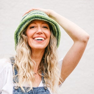 A blonde woman smiles into the camera wearing a green easy bucket hat with a brim, a white T-shirt, and denim overalls. Her hand is on the bucket hat.