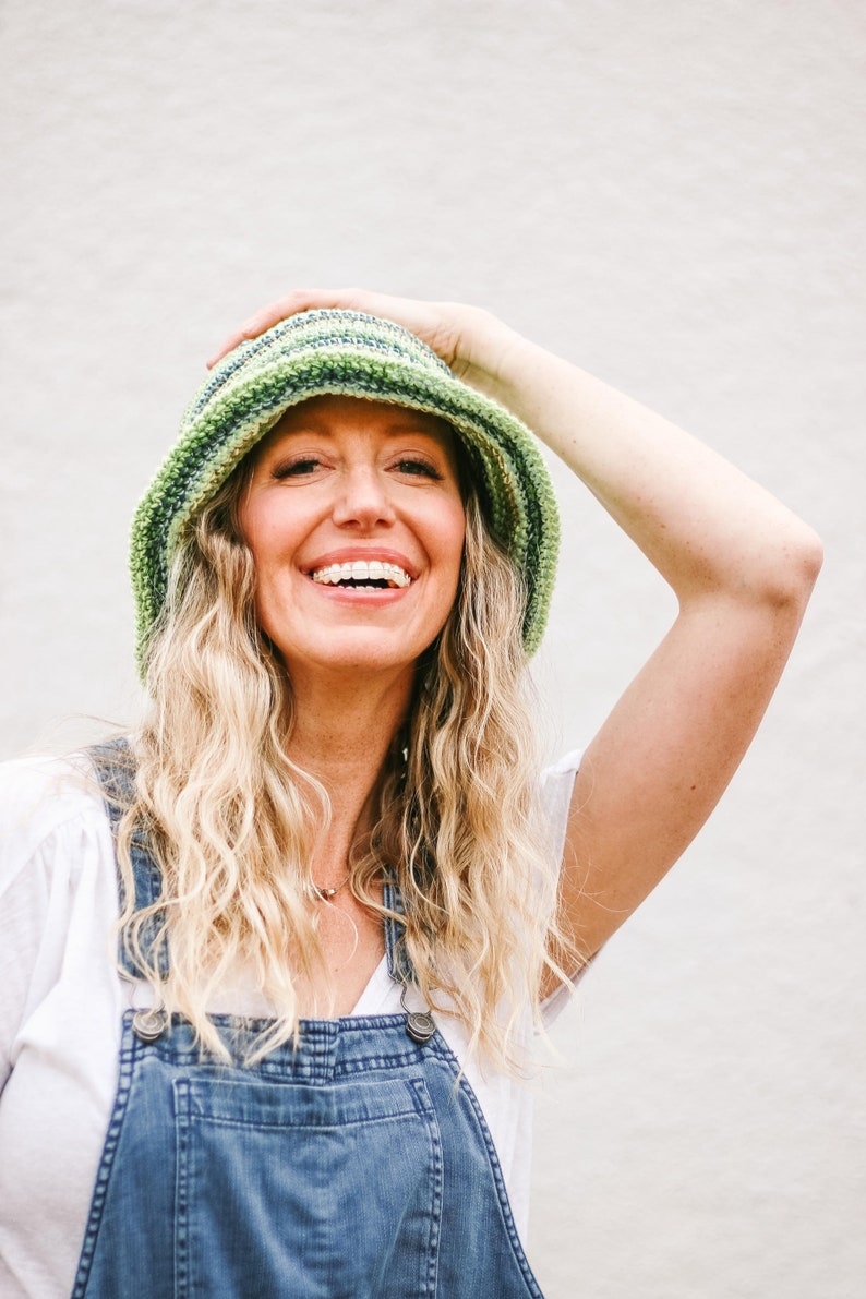 A blonde woman smiles into the camera wearing a green easy bucket hat with a brim, a white T-shirt, and denim overalls. Her hand is on the bucket hat.