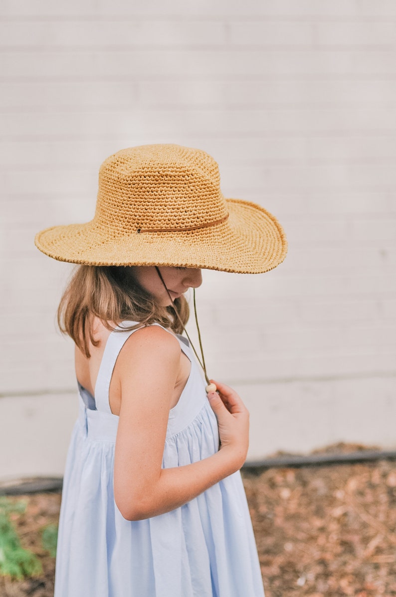 A little girl is looking down, smiling, wearing a brown crochet sun hat and a blue dress. Her hand is on the hat's adjuster.