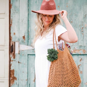 A blonde woman standing in front of a green door, looking down, wearing a sun hat and a white dress, with an farmer's market bag full of vegetables, fruits, and flowers.