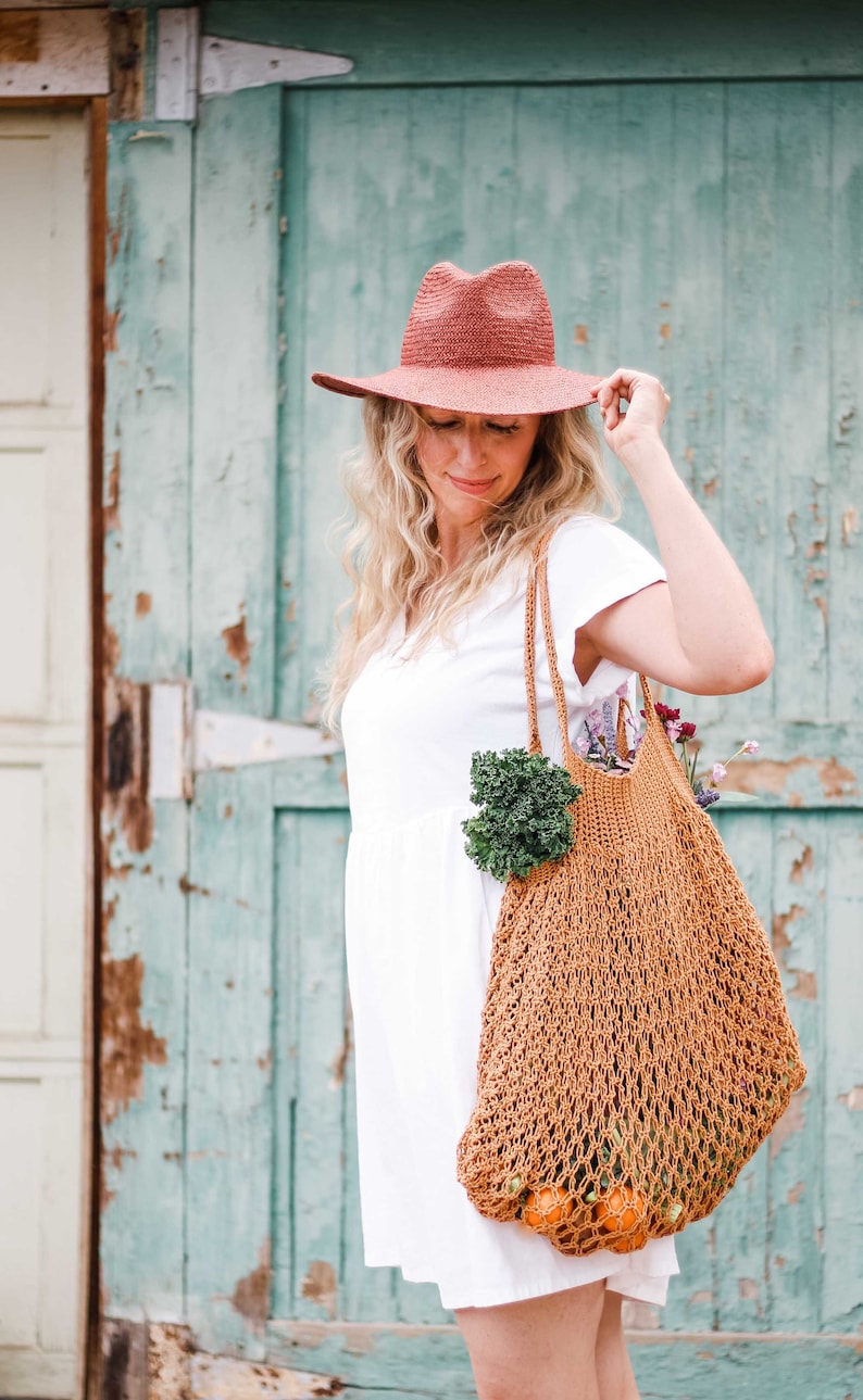 A blonde woman standing in front of a green door, looking down, wearing a sun hat and a white dress, with an farmer's market bag full of vegetables, fruits, and flowers.