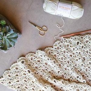 An in-progress crochet summer top featuring the open fan stitch with a crochet hook, a yarn skein, scissors, and some potted plants.