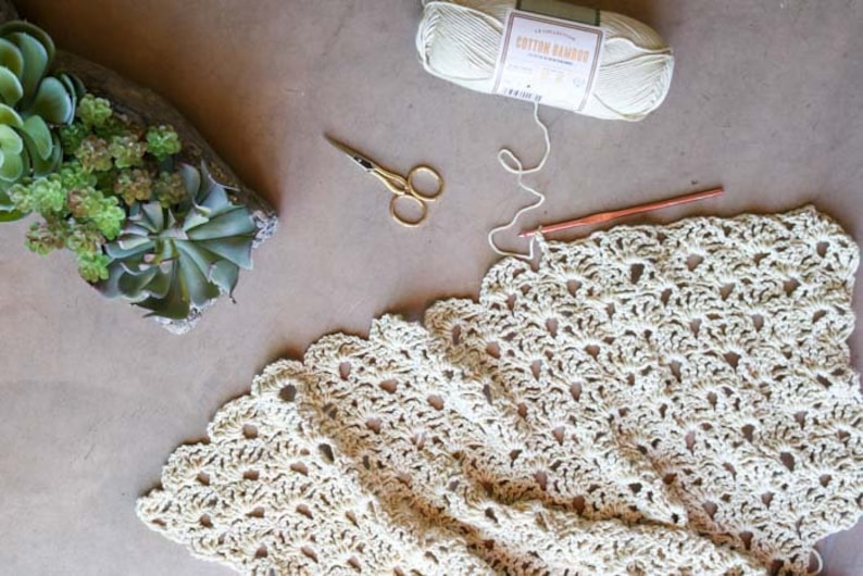An in-progress crochet summer top featuring the open fan stitch with a crochet hook, a yarn skein, scissors, and some potted plants.