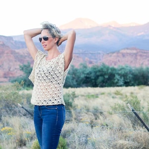 A blonde woman standing on a field fixing her hair wearing a women's boho lace top, a black bandeau and denim jeans.