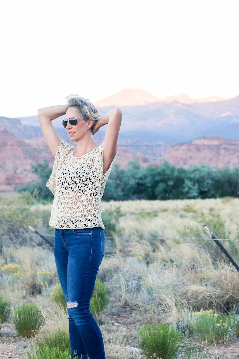 A blonde woman standing on a field fixing her hair wearing a women's boho lace top, a black bandeau and denim jeans.