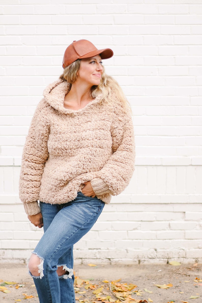 A blonde woman standing, looking sideways, wearing a brown leather baseball cap, a beige sherpa hooded sweatshirt, and ripped denim jeans with her hand on the front pocket of the jeans.