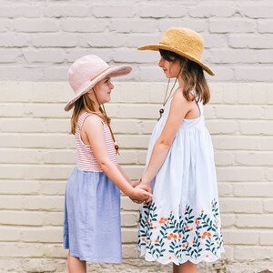 Two little girls face each other, smiling and holding hands, wearing a matching summer hat, with the taller kid wearing a brown one and the other kid wearing a pink one.