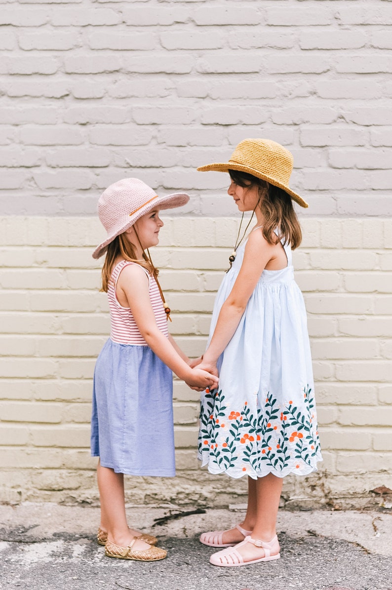 Two little girls face each other, smiling and holding hands, wearing a matching summer hat, with the taller kid wearing a brown one and the other kid wearing a pink one.