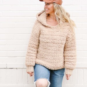 A blonde woman standing with her foot propped on a white wall, smiling, wearing a brown leather baseball cap, a beige sherpa hoodie, and ripped denim jeans.