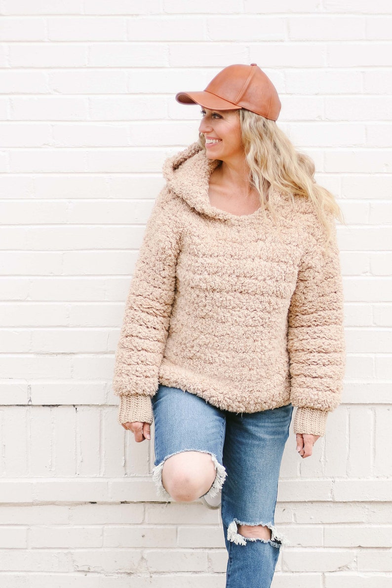 A blonde woman standing with her foot propped on a white wall, smiling, wearing a brown leather baseball cap, a beige sherpa hoodie, and ripped denim jeans.