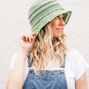 A blonde woman standing in front of a white wall, looking sideways smiling wearing a green gender neutral hat, a white t-shirt, and denim overalls with her hand holding the brim of the hat.