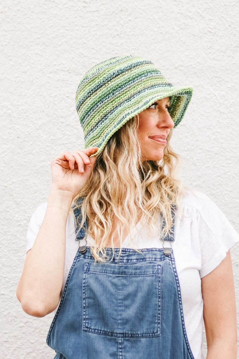 A blonde woman standing in front of a white wall, looking sideways smiling wearing a green gender neutral hat, a white t-shirt, and denim overalls with her hand holding the brim of the hat.