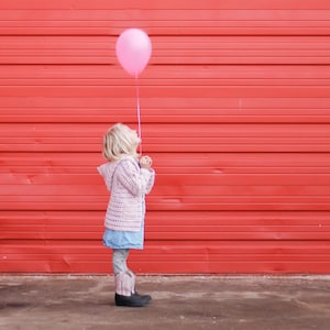 May include: A young person stands in front of a red wall, gazing upwards at a pink balloon. They are wearing a light pink crocheted hooded jacket, a blue denim dress, gray leggings, and pink and black cowboy boots. The balloon is tied to a string.