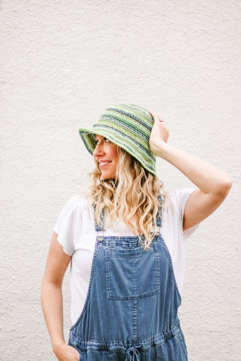A blonde woman standing in front of a white wall, smiling, looking sideways while wearing a green bucket hat, a white t-shirt, and denim overalls with one hand on the hat and the other on the pocket of the overalls.