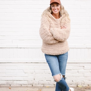A blonde woman standing with her hands and legs crossed, smiling, wearing a brown cap, beige sherpa hooded sweatshirt, ripped denim jeans, and a pair of white sneakers.