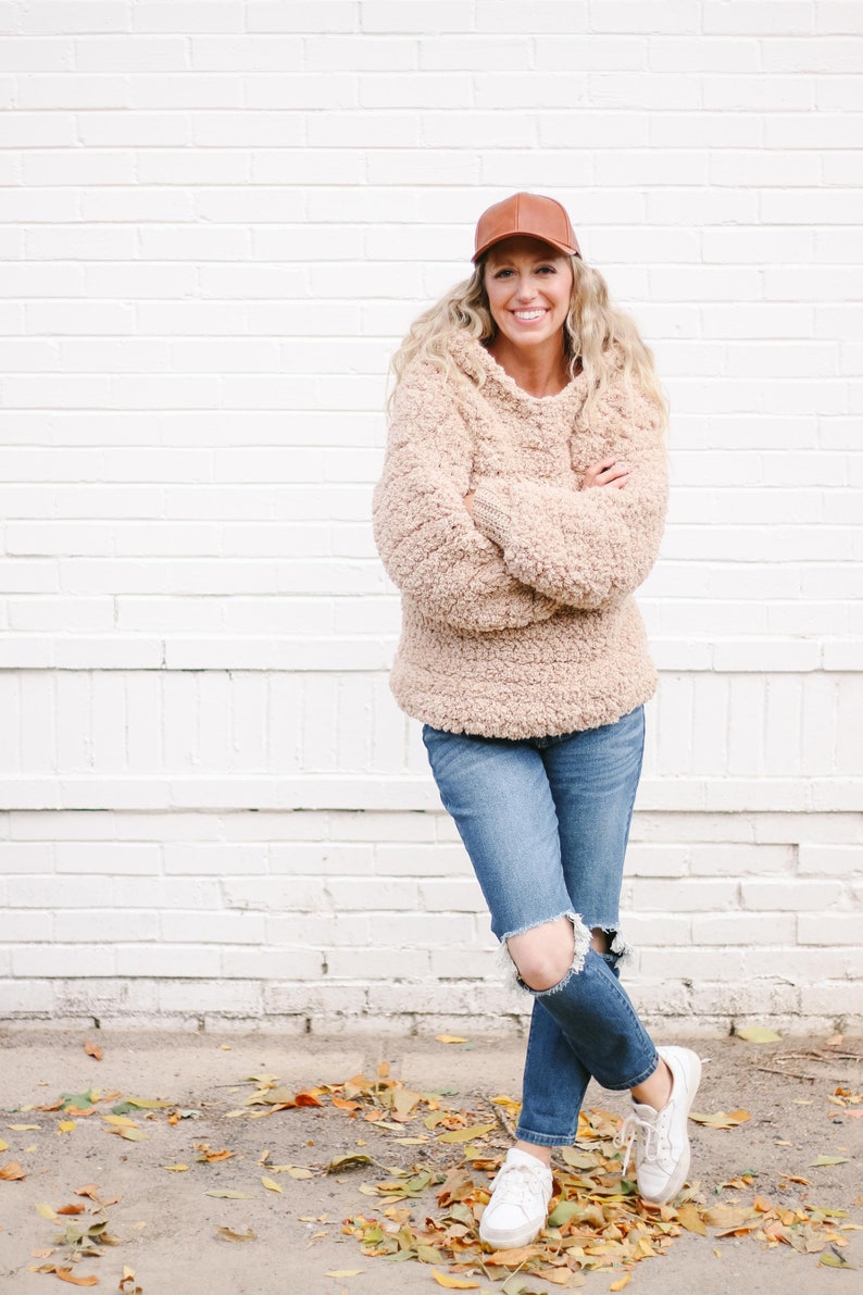 A blonde woman standing with her hands and legs crossed, smiling, wearing a brown cap, beige sherpa hooded sweatshirt, ripped denim jeans, and a pair of white sneakers.