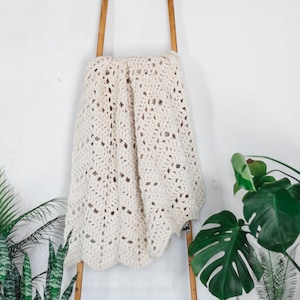 May include: A cream-coloured, crocheted blanket draped over a wooden ladder. The blanket has a scalloped edge and an open, airy design. The ladder is made of wood. Green plants are in the background.