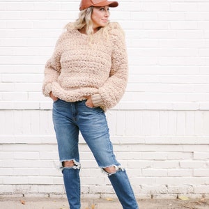 A blonde woman stands in front of a white wall, looking sideways. She is wearing a brown leather ball cap, a beige women's crochet hoodie, denim ripped jeans, and white sneakers. Her hands are on the front pockets of her jeans.
