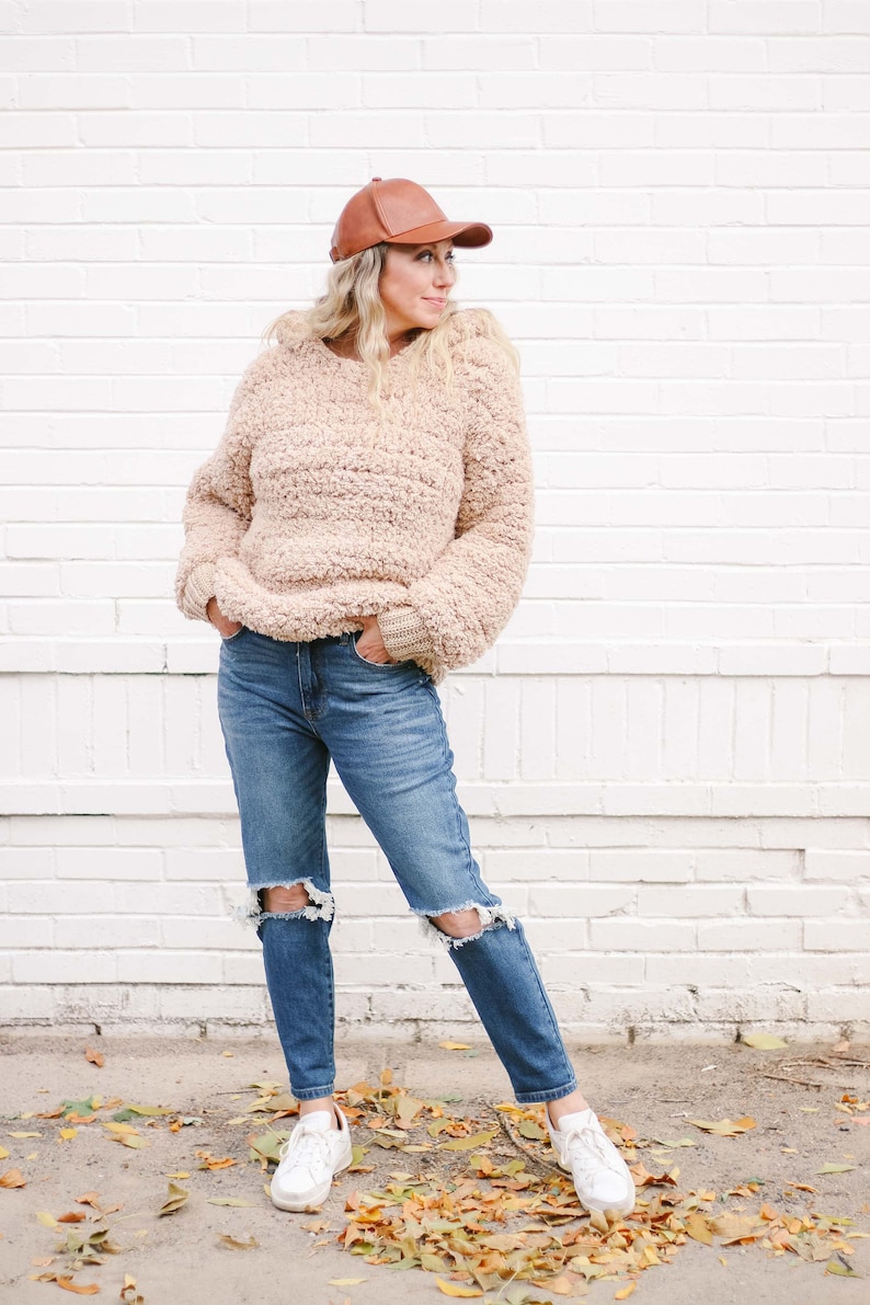A blonde woman stands in front of a white wall, looking sideways. She is wearing a brown leather ball cap, a beige women's crochet hoodie, denim ripped jeans, and white sneakers. Her hands are on the front pockets of her jeans.
