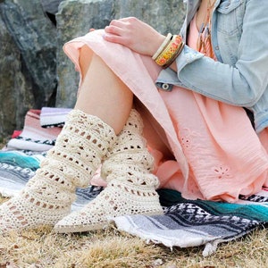 A woman sitting on a mat by the grass wearing a pair of lacy crochet shoes, a pink dress, a denim jacket and some boho accessories.