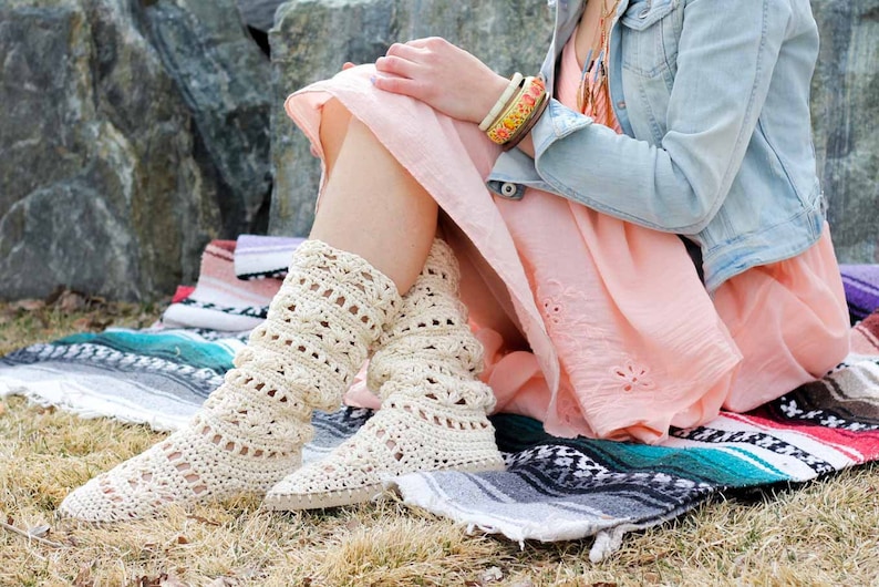 A woman sitting on a mat by the grass wearing a pair of lacy crochet shoes, a pink dress, a denim jacket and some boho accessories.