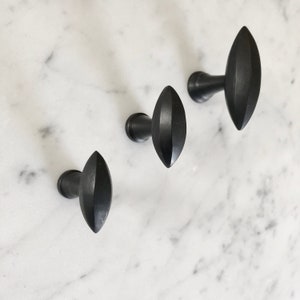 Könnte beinhalten: Drei schwarze Metallhaken mit einem rautenförmigen Design. Die Haken sind auf einer weißen Marmorfläche montiert.