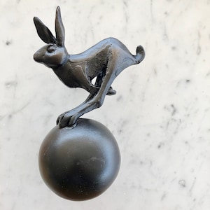 May include: A black metal sculpture of a rabbit leaping onto a black sphere. The sculpture is a decorative piece for a home or office.