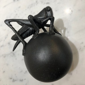 Könnte beinhalten: Eine schwarze Metallskulptur eines Grashüpfers, der auf einer schwarzen Kugel sitzt.