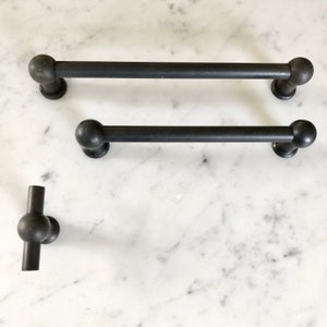 Peut inclure: Trois poignées de tiroir en métal noir avec des extrémités arrondies. Les deux poignées du haut sont longues et droites, tandis que la poignée du bas est en forme de T.