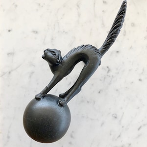 Op de afbeelding: Zwarte keramische kattenfiguur met een opgeheven staart, staand op een zwarte bol. De kat staat in een speelse houding, met zijn kop naar achteren gekanteld en zijn voorpoten omhoog.