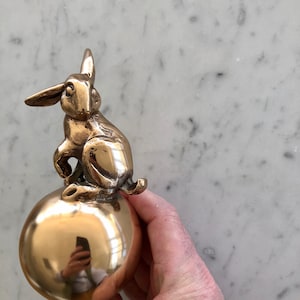 Könnte beinhalten: Eine goldfarbene Hasenfigur, die auf einer runden, goldfarbenen Kugel sitzt. Der Hase hat ein detailliertes, realistisches Design und sitzt aufrecht mit hochgestellten Ohren.