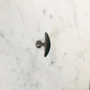 May include: A dark brown metal hook with a crescent-shaped handle. The hook is mounted on a white marble surface.