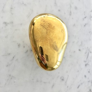 May include: A smooth, polished, golden brass paperweight in the shape of a rounded oval.