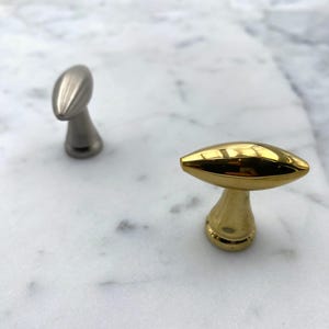 Könnte beinhalten: Zwei Schrankknöpfe, einer silberfarben und einer golden, mit einer abgerundeten, ovalen Oberseite und einer zylindrischen Basis.