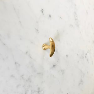 May include: A gold-colored metal hook with a crescent-shaped top. The hook is mounted on a white marble surface.
