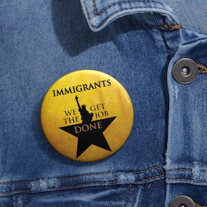 May include: A yellow button with a black star and the text "Immigrants We Get The Job Done". The star has a silhouette of the Statue of Liberty inside.
